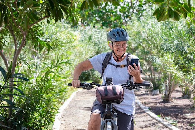 Active smiling senior woman cycling with her electro bike in a green park wearing helmet Mature attractive woman stops using cellphone reading message Retirement and healthy lifestyle concept