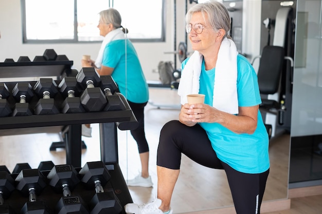 Active smiling senior woman drinks water glass doing exercises in the gym to stay fit Sport concep