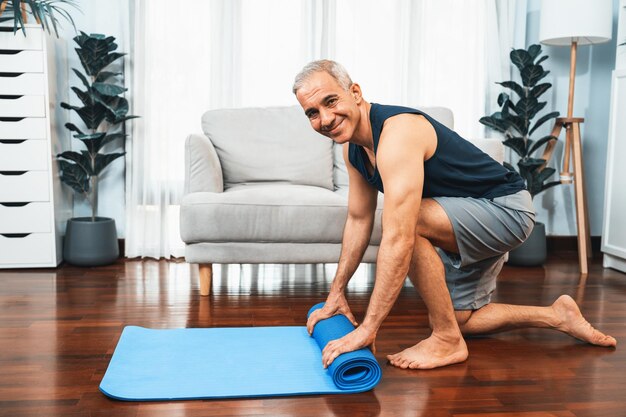 Active and sporty senior man preparing rolling fitness exercising mat on living room floor at home home exercise as concept of healthy fit body lifestyle after retirement clout