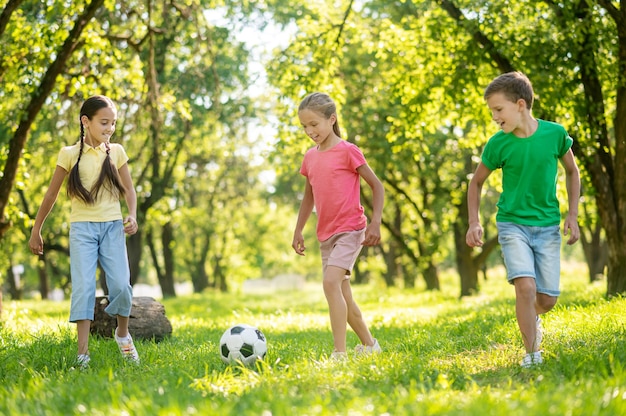 Active summer. Blonde boy and two long-haired cheerful girlfriends playing football on green lawn on sunny day