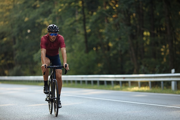 Active young cyclist in black helmet, protective glasses and sport outfit riding bike on paved road.