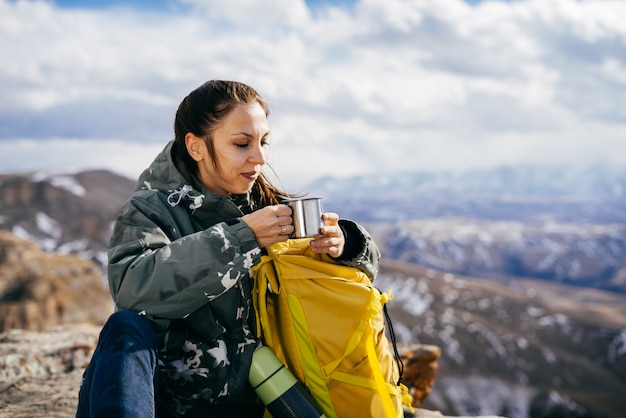 An active young girl in a warm jacket travels through the mountains, enjoys nature, drinks hot tea from a thermos