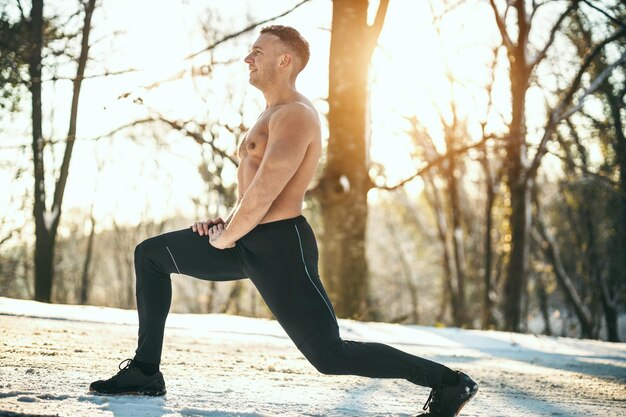 An active young man is doing stretching exercises in the forest road during the winter training outside.