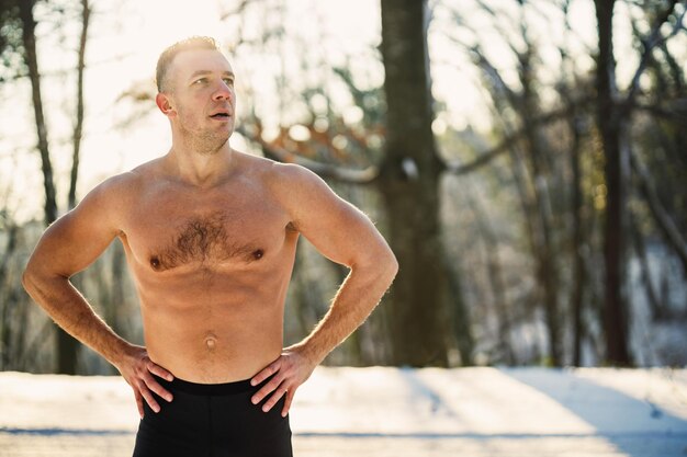 An active young man is doing stretching exercises in the forest during training outside in the winter snowy day.