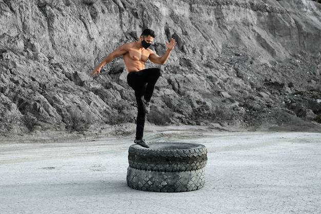 Active young man with bare torso wearing black protective mask, using heavy tyres for workout at sand quarry. Concept of strength and endurance.