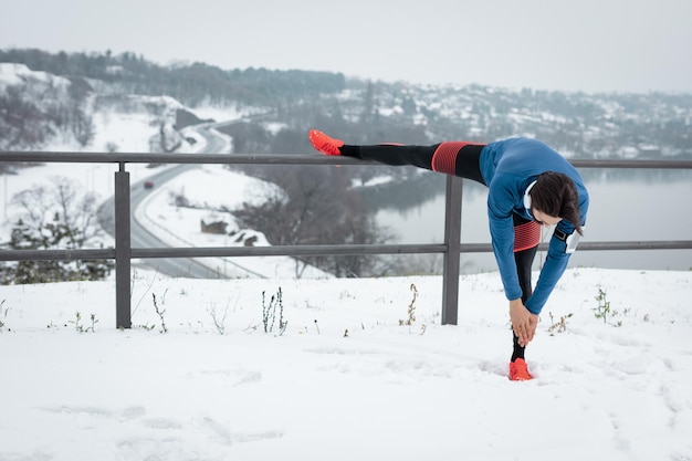 Фото Активный молодой человек с наушниками на ушах растягивается и делает упражнения в общественном месте во время зимних тренировок на улице, пока идет снег. скопируйте пространство.