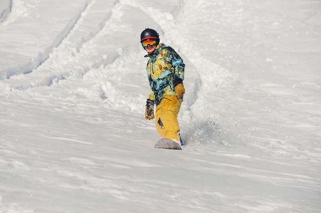 明るい冬の日に雪の丘を下って乗って明るいスポーツウェアでアクティブな若いスノーボーダー