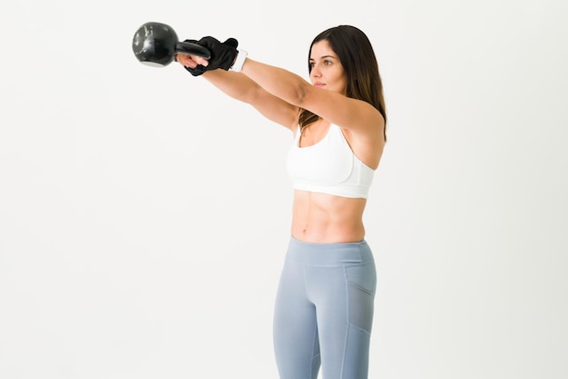 Active young woman in sports clothing training with a kettlebell. Caucasian strong woman with gloves lifting weights against a white background