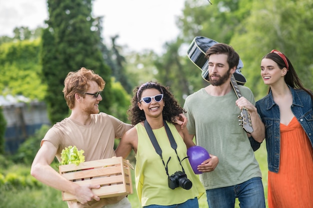 Активность, лето. Молодые радостные обнявшиеся парни с гитарой и едой и девушки с фотоаппаратом и мячом собираются на пикник на природе