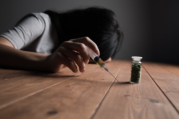 The addict holds a syringe and pills in a vessel on the table