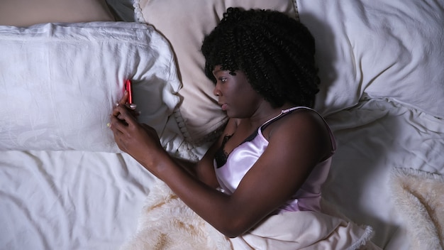 Пристрастившаяся к интернету серьезная темнокожая дама смотрит на современный красный смартфон и смотрит на утреннюю кровать, лежащую на кровати размера «queen-size» в спальне.