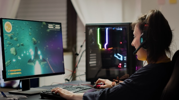Зависимая женщина играет в космический шутер с наушниками, беспроводным джойстиком и мощным персональным компьютером с rgb-подсветкой. кибер-геймер с гарнитурой и джойстиком играет в виртуальные игры на чемпионате по играм