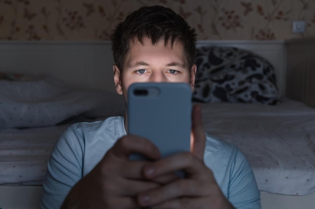 Photo addicted man sitting near bed and surfing on the internet with smartphone social media dependency