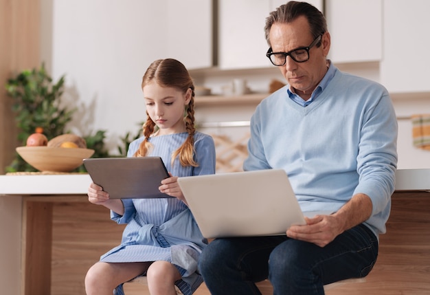 Uomo intelligente in pensione dipendente seduto a casa e utilizzando gadget digitali con la nipote durante la navigazione in internet