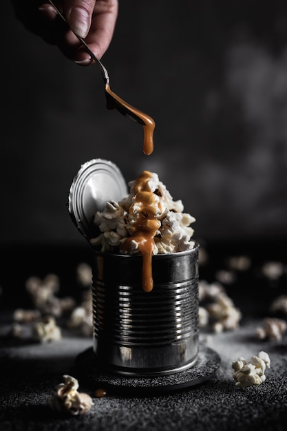 Adding caramel on top of salted popcorn in a steel tin on dark background, moody photo