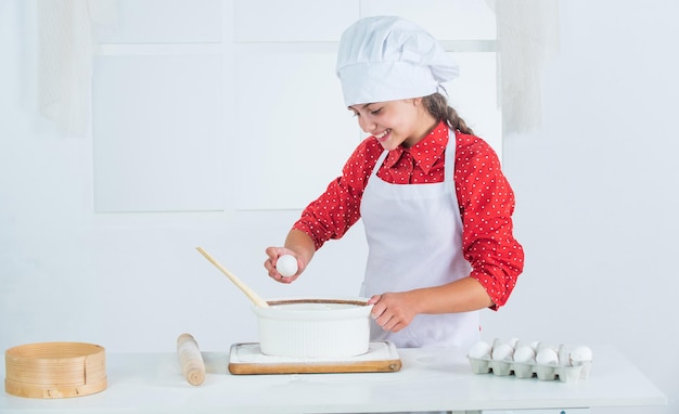 卵を追加します。食事の時間。キッチンで料理をする幸せな子。キッチンでクッキーを焼く。プロの熟練したパン屋。シェフの制服と帽子の子供。生地を準備している十代の少女。レシピでケーキを作る。