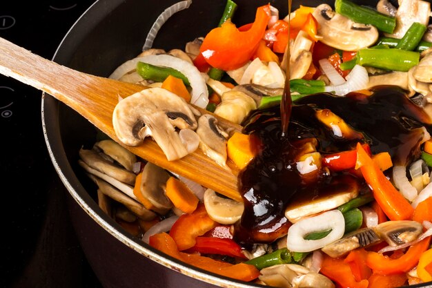 Adding sauce to fried vegetables with mushrooms in a frying pan on a dark background