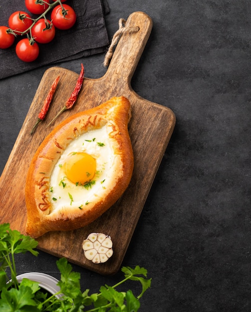 Adjarian khachapuri dish Traditional Georgian food made from homemade bread with cheese and egg yolk on a wooden board with tomatoes and fresh herbal Top view and copy space