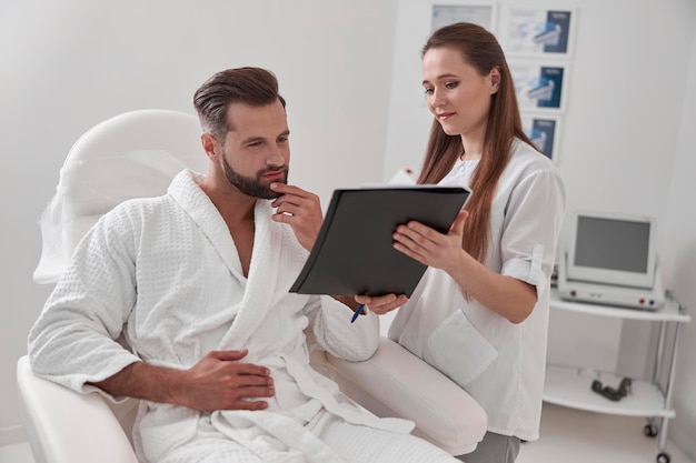 Administrator shows clipboard with form to client sitting in armchair in cosmetological clinic