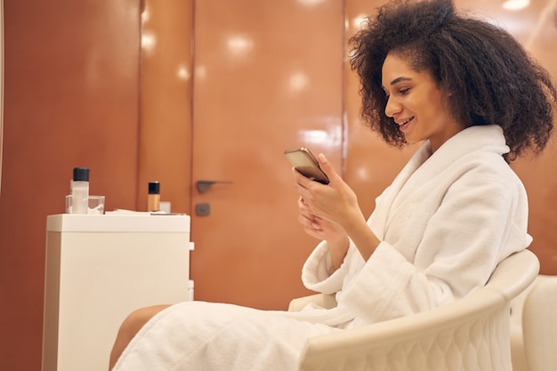 Admirable curly lady holding a smartphone and checking messages with curiosity while resting in armchair of spa salon