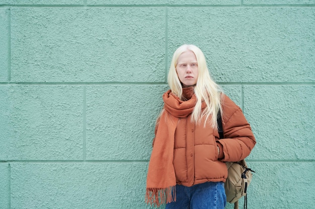 Adolescent albino girl in jeans jacket and scarf standing by blue wall