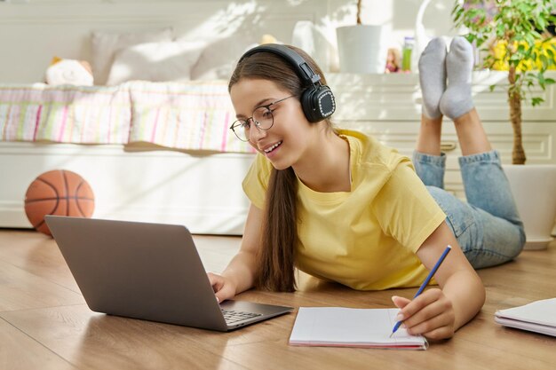 Adolescent girl in headphones using laptop lying on floor writing in notebook