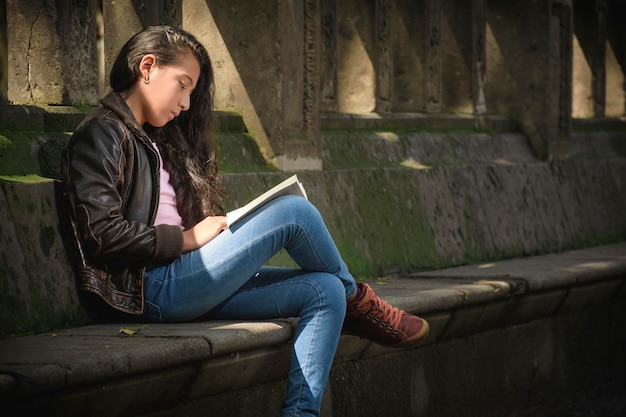 Adolescent girl is very attentive reading a book she is sitting on a bench in the park it is a beautiful day and she enjoys reading