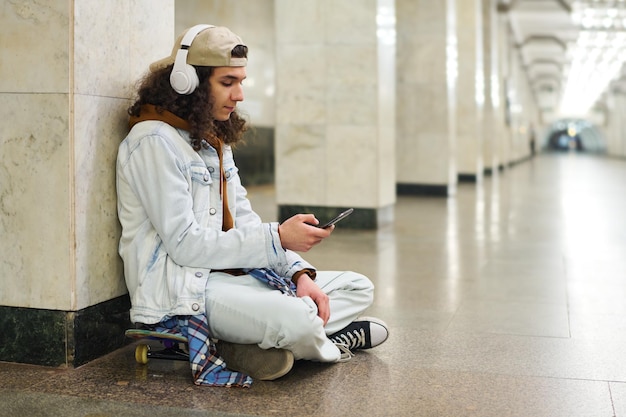 Adolescent guy in casualwear sitting on skateboard by white marble column