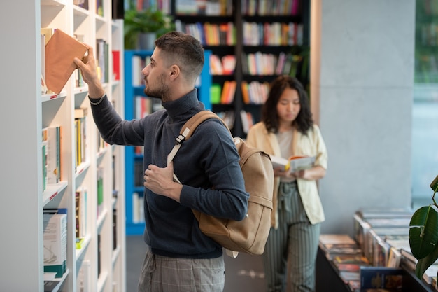 Adolescente man in vrijetijdskleding die boek van boekenplank neemt