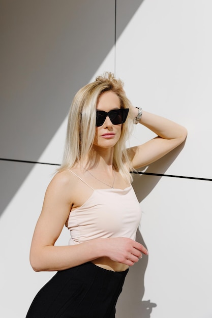 Adorabele blonde volwassen vrouw in top en broek die bij de witte muur staat en naar de camera kijkt