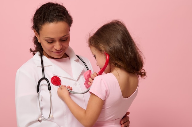 Adorable 4 y.o. child girl playing doctor, using stethoscope, listening to chest and lungs breathing of doctor with pink ribbon on chest. Concept of educational program for supporting cancer patients