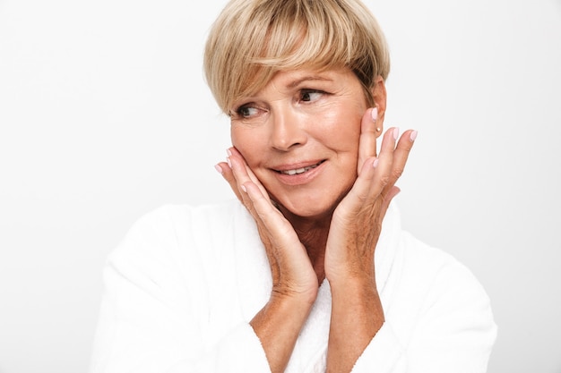  adorable adult woman with short blond hair wearing white housecoat touching her face isolated over white wall