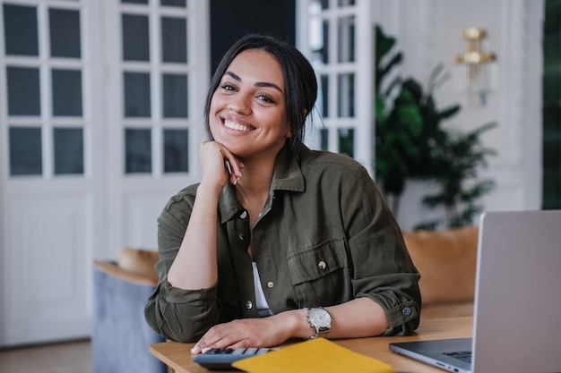 Adorable African American girl in casual shirt sitting at desk with laptop looks at camera