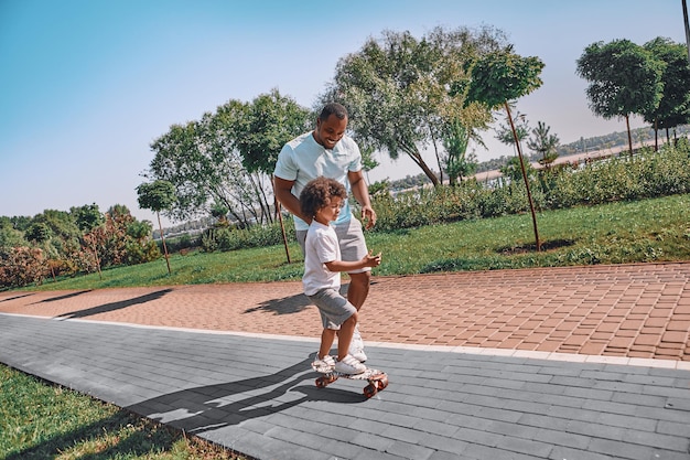 Adorable AfroAmerican kid skateboarding with a help of his caring father