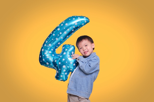 Adorable Asian four year old boy celebrating his birthday holding number 4 blue balloon on orange colored background with clipping path