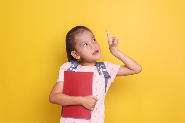 Adorable asian girl student holding book and pointing up to empty space isolated on yellow backgroun