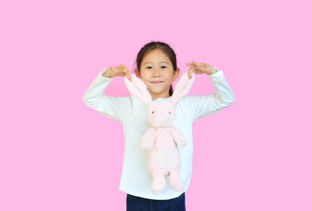 Adorable asian kid girl holding ears of pink rabbit doll isolated on pink background Happy child on Easter