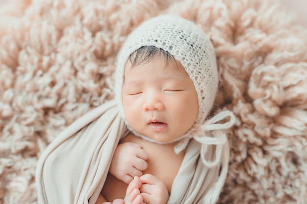 毛皮で寝ている繭のニット帽とwrapprdを持つ愛らしいアジアの生まれたばかりの赤ちゃん