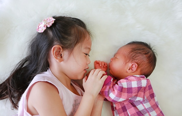 Adorable Asian older sister and newborn baby lying on white fur background.