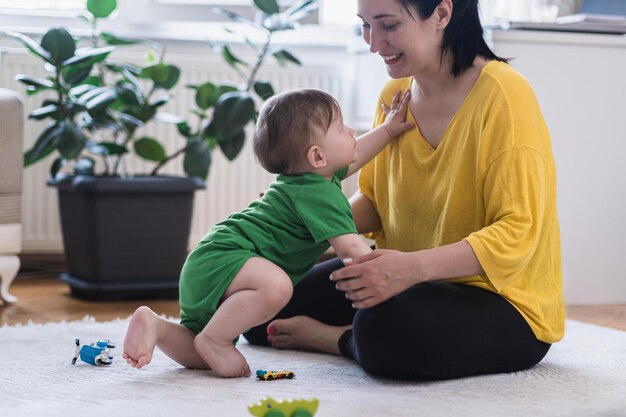 写真 散らばったおもちゃの近くの白い敷物の上に座っているママに這う愛らしい男の子