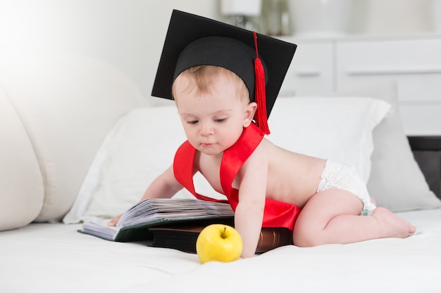 Adorable baby boy in graduation cap posing with apple and big book