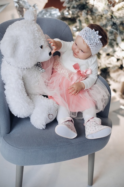 Adorable baby girl with teddy bear on armchair by Christmas tree.