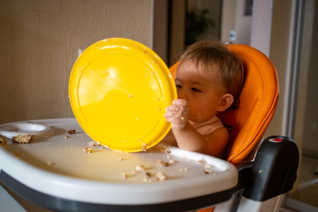 愛らしい赤ちゃんがテーブルでプラスチックプレートで遊んでいます。小さな子供は食べた後、赤ちゃんの椅子にふける