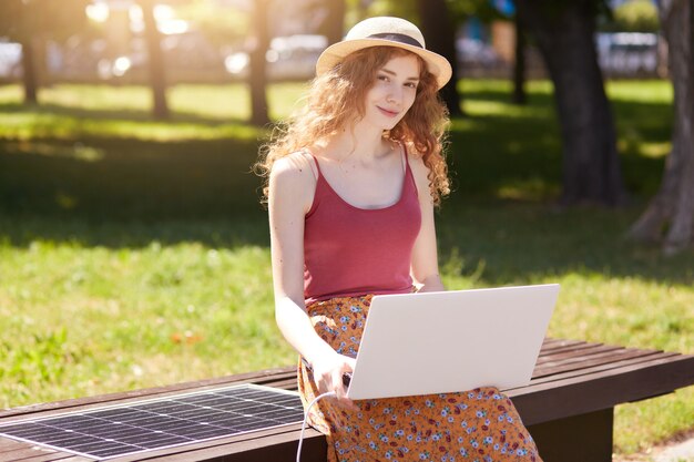 Adorable beautiful female sitting on park wooden bench, using her laptop, wearing straw hat