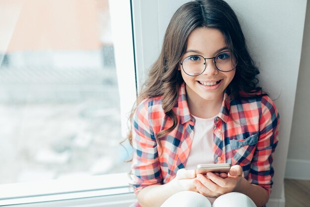 窓の近くの壁と笑顔のウェブサイトのバナーのそばにスマートフォンで座っている愛らしい眼鏡をかけた女の子