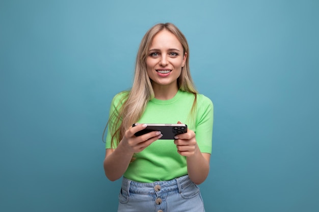 Adorable blond girl in casual outfit plays a game on the phone on a blue background