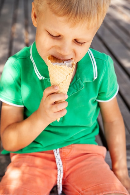 Adorable blond haired little boy in fashionable sunglasses biting yummy vanilla ice cream cone while spending summer day in park