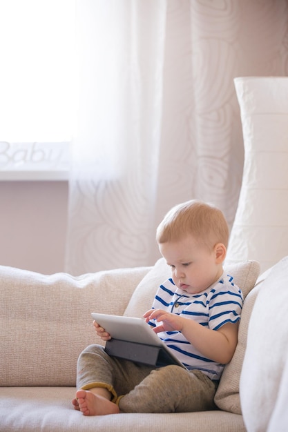 Adorable blond toddler boy laying on the sofa and playing with tablet pc at home indoors