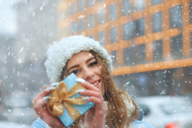 사랑스러운 금발 여성은 파란 선물 상자를 들고 눈이 내리는 동안 도시를 걷고 있는 흰색 니트 모자를 쓰고 있습니다.