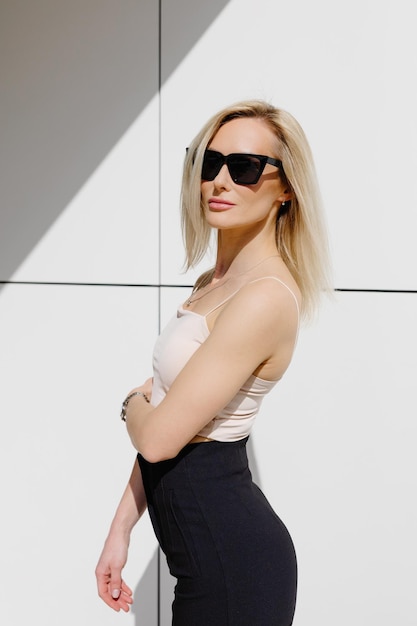 Adorable blonde adult woman wearing in top and pants standing at white wall looking at camera